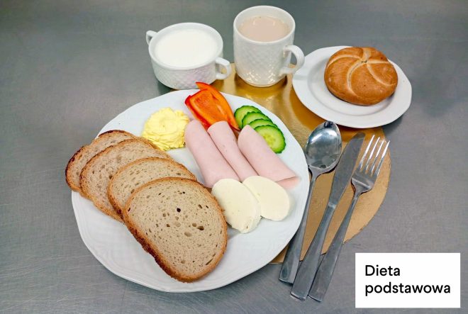 Śniadanie w diecie podstawowej: na talerzu kromki chleba, plasterki szynki, biały ser, masło oraz warzywa – papryka i ogórek. Obok stoją kubki z mlekiem i napojem oraz bułka na talerzyku. W prawym dolnym rogu napis „Dieta podstawowa”