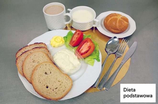 Śniadanie w diecie podstawowej: na talerzu kromki chleba, jogurt naturalny w pojemniku, masło, sałata i pomidor. Obok znajduje się kubek z napojem, miseczka z mlekiem oraz bułka na osobnym talerzyku. W prawym dolnym rogu napis „Dieta podstawowa”