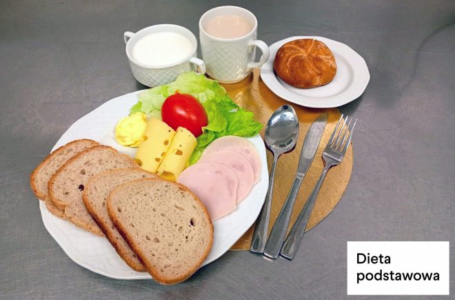 Śniadanie w diecie podstawowej: na talerzu kromki chleba, plasterki szynki, żółty ser, masło, sałata i pomidor. Obok znajduje się bułka na osobnym talerzyku oraz kubek z napojem i miseczka z mlekiem. W prawym dolnym rogu napis „Dieta podstawowa”