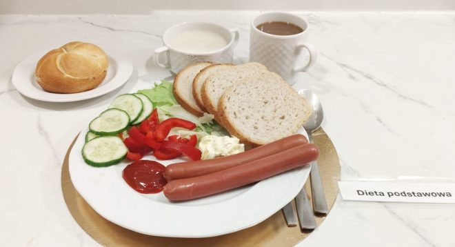 Śniadanie w diecie podstawowej: na talerzu parówki, kromki białego pieczywa, sałata, plasterki ogórka, papryka, porcja twarogu oraz ketchup. W tle bułka, kubek z mlekiem i kubek z kawą. Posiłek podany na tacy