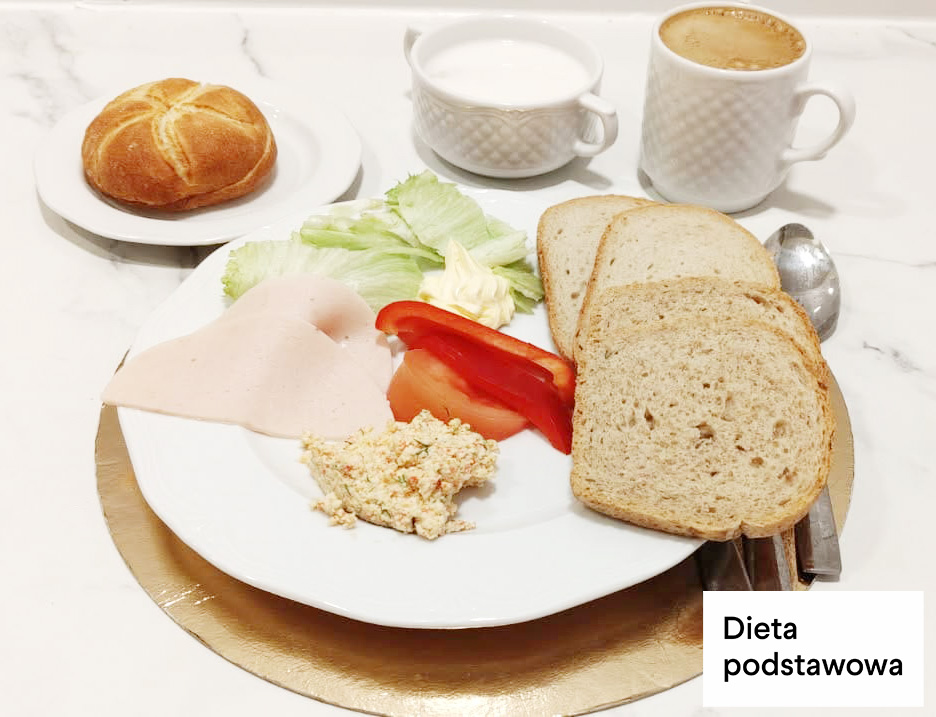 Śniadanie w diecie podstawowej podane na dużym białym talerzu. Na talerzu znajdują się plastry jasnej wędliny drobiowej, liście sałaty, kawałki czerwonej papryki oraz porcja twarożku z dodatkiem ziół. Obok leżą dwie kromki chleba pszenno-żytniego. W tle widoczna jest bułka kajzerka na osobnym talerzyku oraz dwa białe kubki – jeden z mlekiem, drugi z kawą. W prawym dolnym rogu zdjęcia napis „Dieta podstawowa”