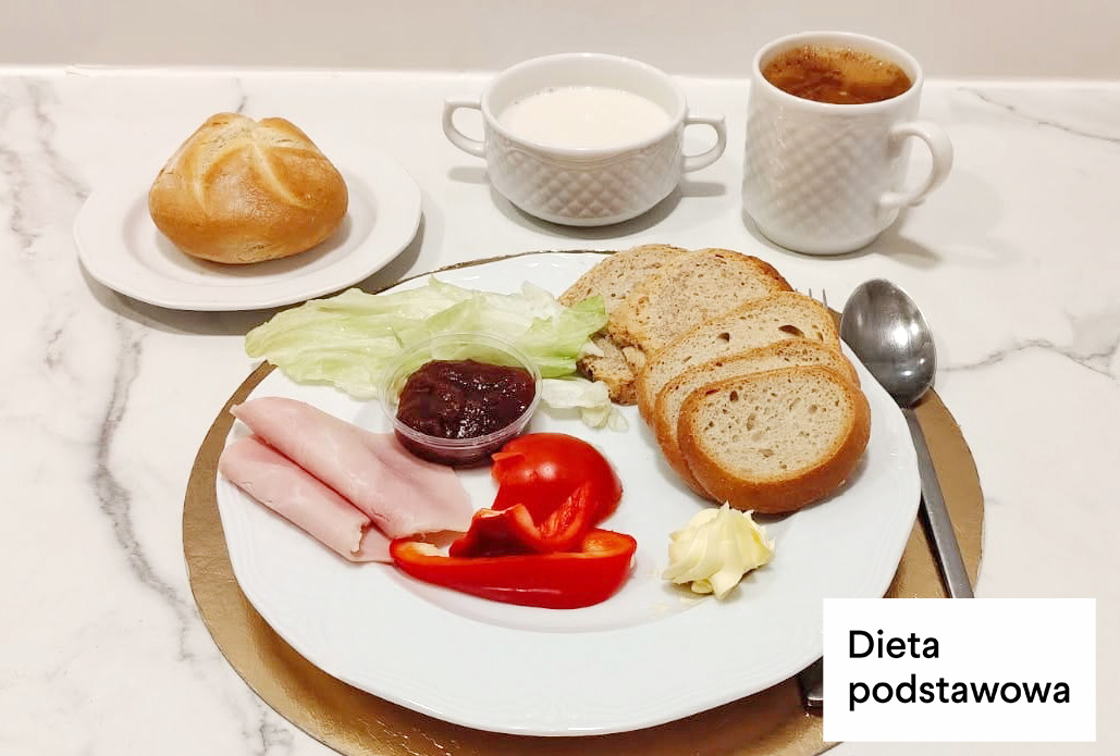 Na talerzu dwie kromki chleba, plaster szynki drobiowej, porcja masła, liście sałaty, czerwona papryka oraz mały pojemnik z dżemem. Obok bułka kajzerka na osobnym talerzyku, filiżanka kawy z mlekiem i kubek mleka