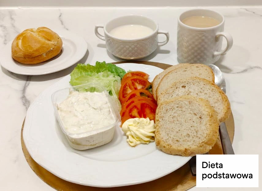 Dwie kromki chleba, twarożek w plastikowym pojemniku, plasterki pomidora, liść sałaty oraz porcja masła. Obok bułka kajzerka, filiżanka kawy z mlekiem i kubek mleka