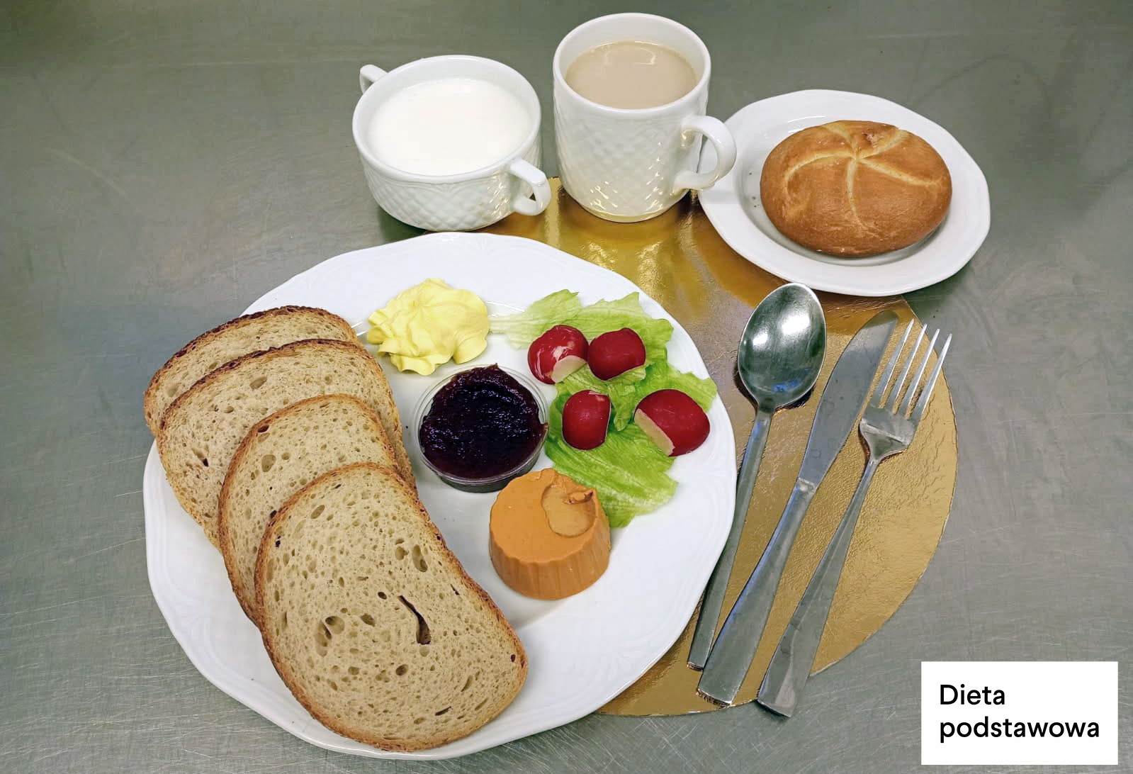 Na talerzu trzy kromki chleba, porcja masła, pasztet drobiowy w krążku, buraczki w małym pojemniku, liście sałaty oraz kilka rzodkiewek. Obok bułka kajzerka, kubek kawy z mlekiem i filiżanka mleka