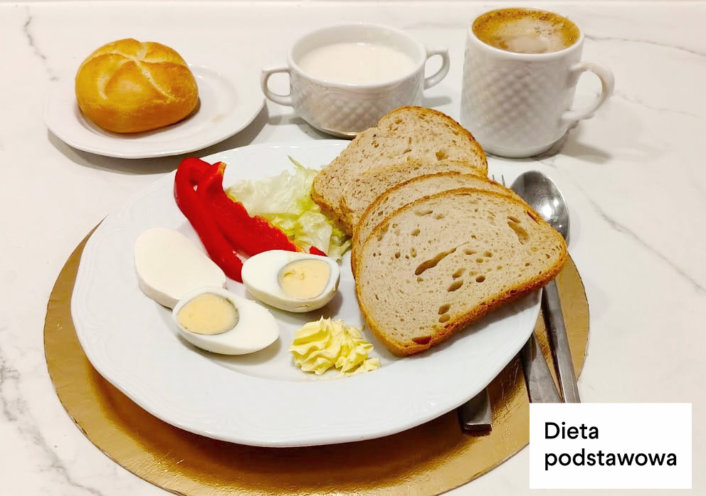 Na białym talerzu dwie kromki chleba pszenno-żytniego, jajko gotowane na twardo przekrojone na połówki, plaster czerwonej papryki, liście sałaty oraz porcja masła. Obok bułka kajzerka na osobnym talerzyku, filiżanka kawy z mlekiem i kubek mleka. Sztućce ułożone po prawej stronie talerza