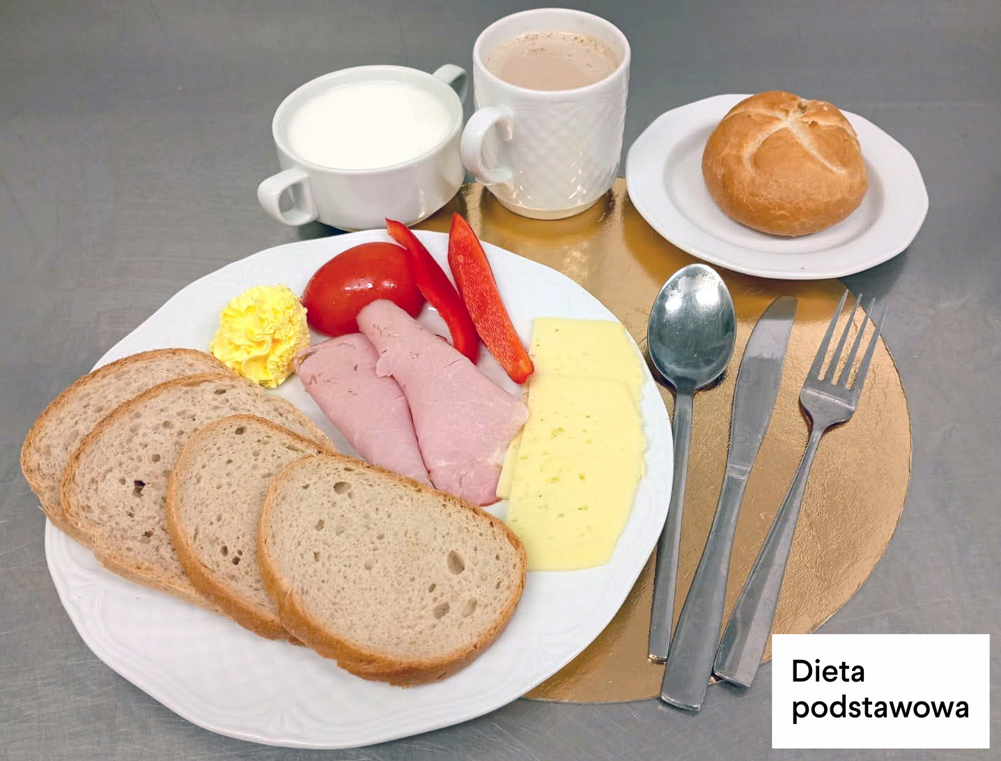 Na talerzu trzy kromki chleba pszenno-żytniego, dwa plasterki sera żółtego, dwa plasterki wędliny drobiowej, porcja masła, kawałki świeżego pomidora oraz paski czerwonej papryki. Obok bułka kajzerka na małym talerzyku. W tle filiżanka mleka oraz kubek kawy z mlekiem. Nakrycie ustawione na złotej podkładce, sztućce po prawej stronie