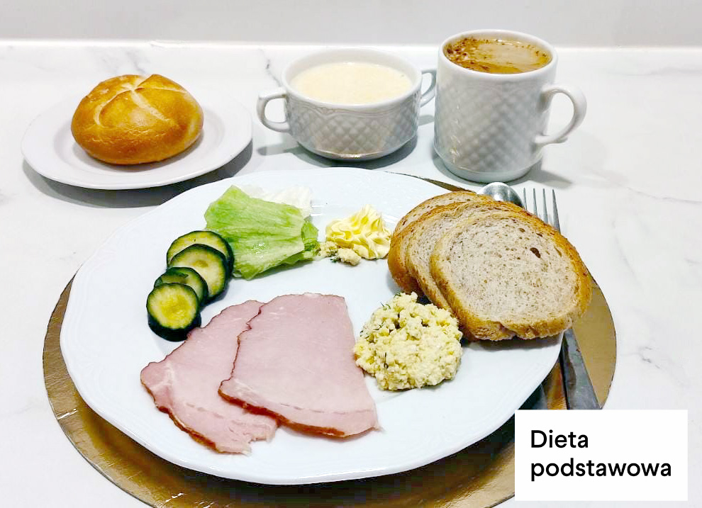 Na białym talerzu trzy plasterki szynki, pasta jajeczna, dwie kromki chleba pszenno-żytniego, liść sałaty oraz kilka plasterków świeżego ogórka. Obok porcja masła. Na osobnym talerzyku bułka kajzerka. W tle filiżanka mleka oraz kubek kawy z mlekiem. Sztućce ułożone po prawej stronie talerza