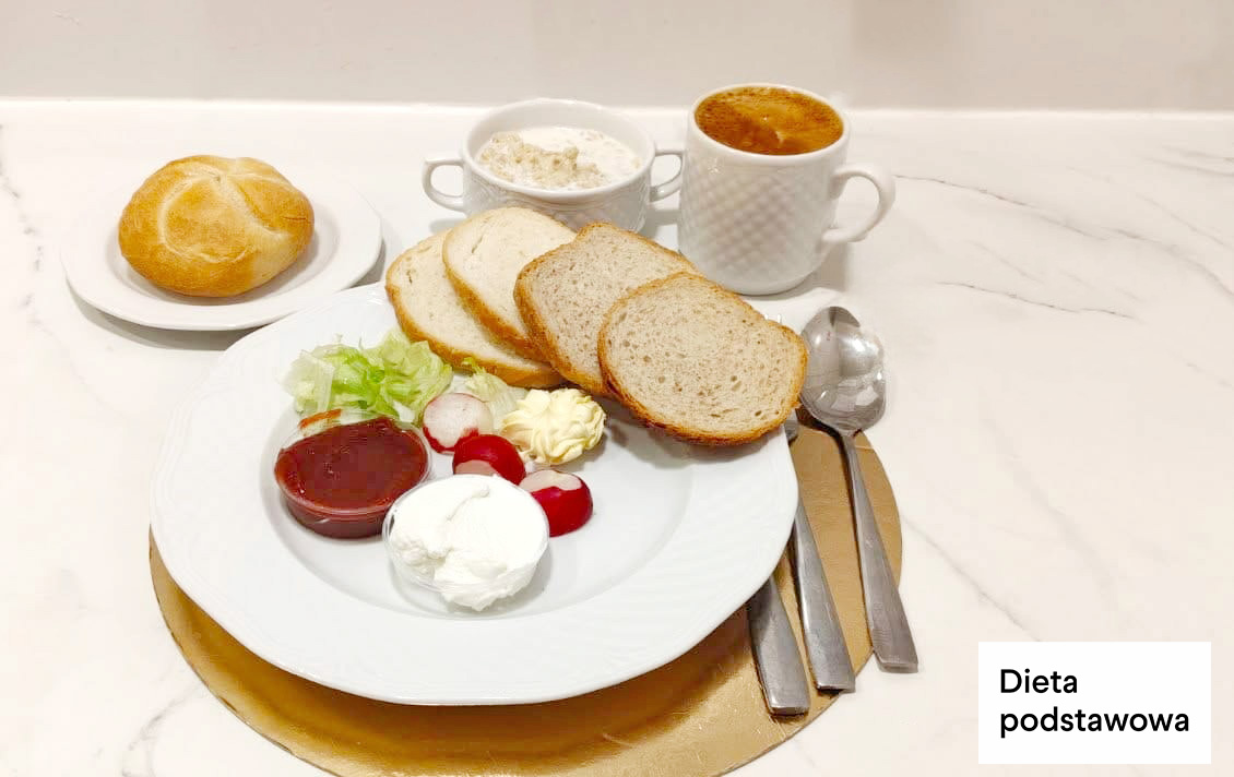 Na talerzu kilka kromek chleba, twarożek, porcja dżemu, sałata i rzodkiewki oraz masło. W tle bułka, kubek kawy zbożowej oraz miseczka z białym serem. Posiłek oznaczony jako dieta podstawowa