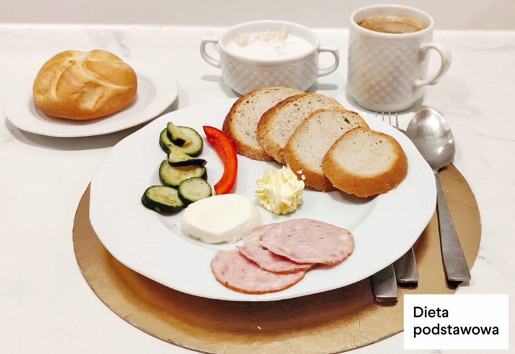 Na talerzu kromki chleba, plasterki wędliny, ser biały, masło oraz świeże warzywa – ogórek i papryka. W tle bułka, miseczka z twarożkiem i kubek kawy zbożowej. Dieta podstawowa