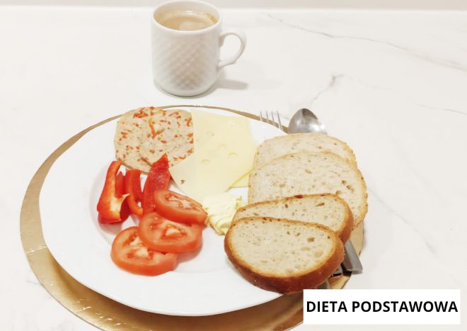 Śniadanie w diecie podstawowej podane na białym talerzu na złotej podkładce. Na talerzu znajdują się kromki pieczywa, plaster żółtego sera, porcja masła oraz wędlina drobiowa. Obok ułożono plasterki czerwonej papryki i pomidora. W tle stoi biały kubek z kawą lub herbatą. W prawym dolnym rogu widoczny napis ‘DIETA PODSTAWOWA’