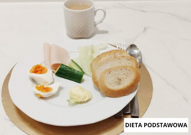 Śniadanie w diecie podstawowej podane na białym talerzu na złotej podkładce. Na talerzu znajdują się dwie kromki pieczywa, przekrojone jajko na miękko, plasterki wędliny oraz kawałek ogórka i liście sałaty. Obok widoczna jest porcja masła. W tle stoi biały kubek z jasnym napojem. Po prawej stronie talerza ułożone są sztućce. W prawym dolnym rogu znajduje się napis ‘DIETA PODSTAWOWA’