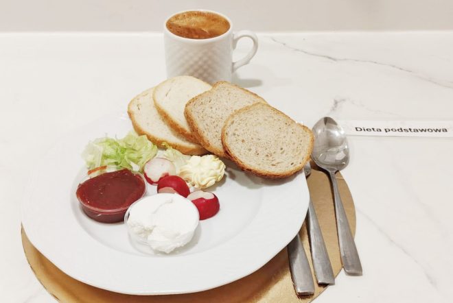 Śniadanie w diecie podstawowej: na białym talerzu kromki pieczywa pszennego, porcja twarogu, rzodkiewki, sałata, porcja masła oraz sos pomidorowy w małym pojemniku. W tle biały kubek z gorącym napojem. Sztućce ułożone po prawej stronie talerza