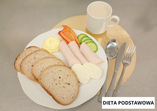 Śniadanie w diecie podstawowej: na białym talerzu kromki pieczywa pszennego, plastry wędliny drobiowej, plasterki jajka na twardo, ogórek, papryka oraz porcja masła. W tle biały kubek z jasnym napojem. Sztućce ułożone obok talerza