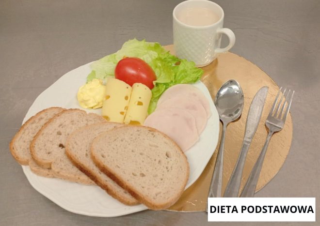 Śniadanie w diecie podstawowej: na białym talerzu cztery kromki pieczywa pszennego, plastry żółtego sera, plasterki wędliny drobiowej, liść sałaty oraz pomidor. Obok porcja masła. W tle biały kubek z jasnym napojem. Sztućce ułożone po prawej stronie talerza