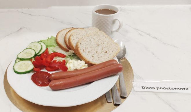 Talerz z posiłkiem w ramach diety podstawowej: dwie parówki, kromki białego pieczywa, plasterki ogórka i papryki oraz sałata. Obok znajduje się ketchup, sztućce oraz kubek z gorącym napojem. Na stole widoczna kartka z napisem „Dieta podstawowa”