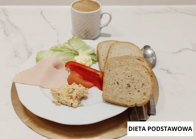 Śniadanie diety podstawowej na białym talerzu: dwie kromki chleba, plaster wędliny drobiowej, porcja twarożku z warzywami, liście sałaty, kawałki pomidora i czerwonej papryki oraz porcja masła. W tle kubek kawy, obok sztućce