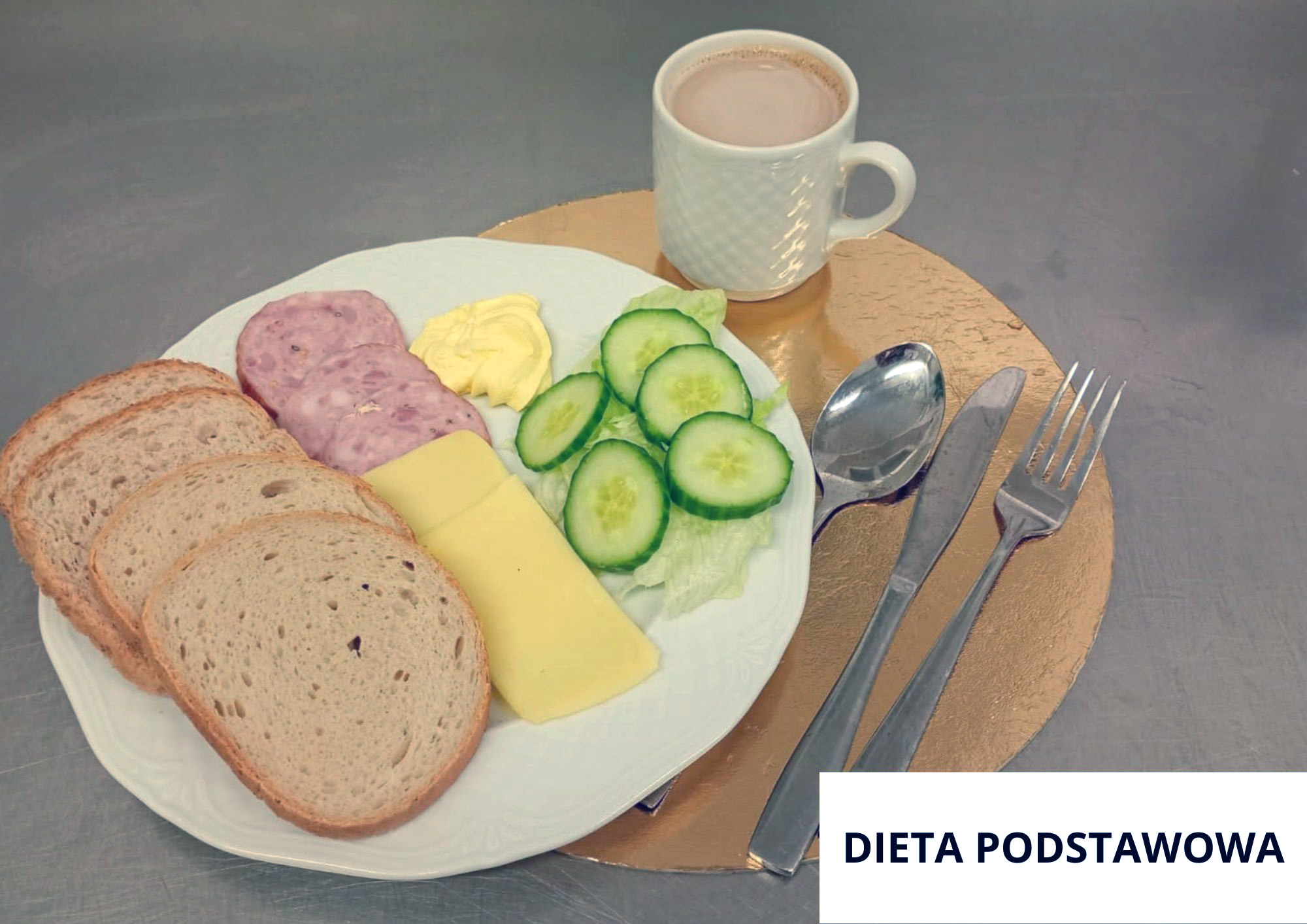 Na stalowym blacie, na okrągłej złotej podkładce stoi biały talerz z trzema kromkami pieczywa, plastrem żółtego sera, dwoma plasterkami wędliny, listkiem sałaty i kilkoma plasterkami świeżego ogórka ułożonymi w wachlarz. Obok ogórka widać małą porcję masła. Z tyłu, na tej samej podkładce, stoi biały kubek z jasnym napojem (kawa z mlekiem lub kakao). Po prawej stronie talerza leżą sztućce: łyżka, nóż i widelec. W prawym dolnym rogu zdjęcia znajduje się biała plansza z napisem „DIETA PODSTAWOWA”