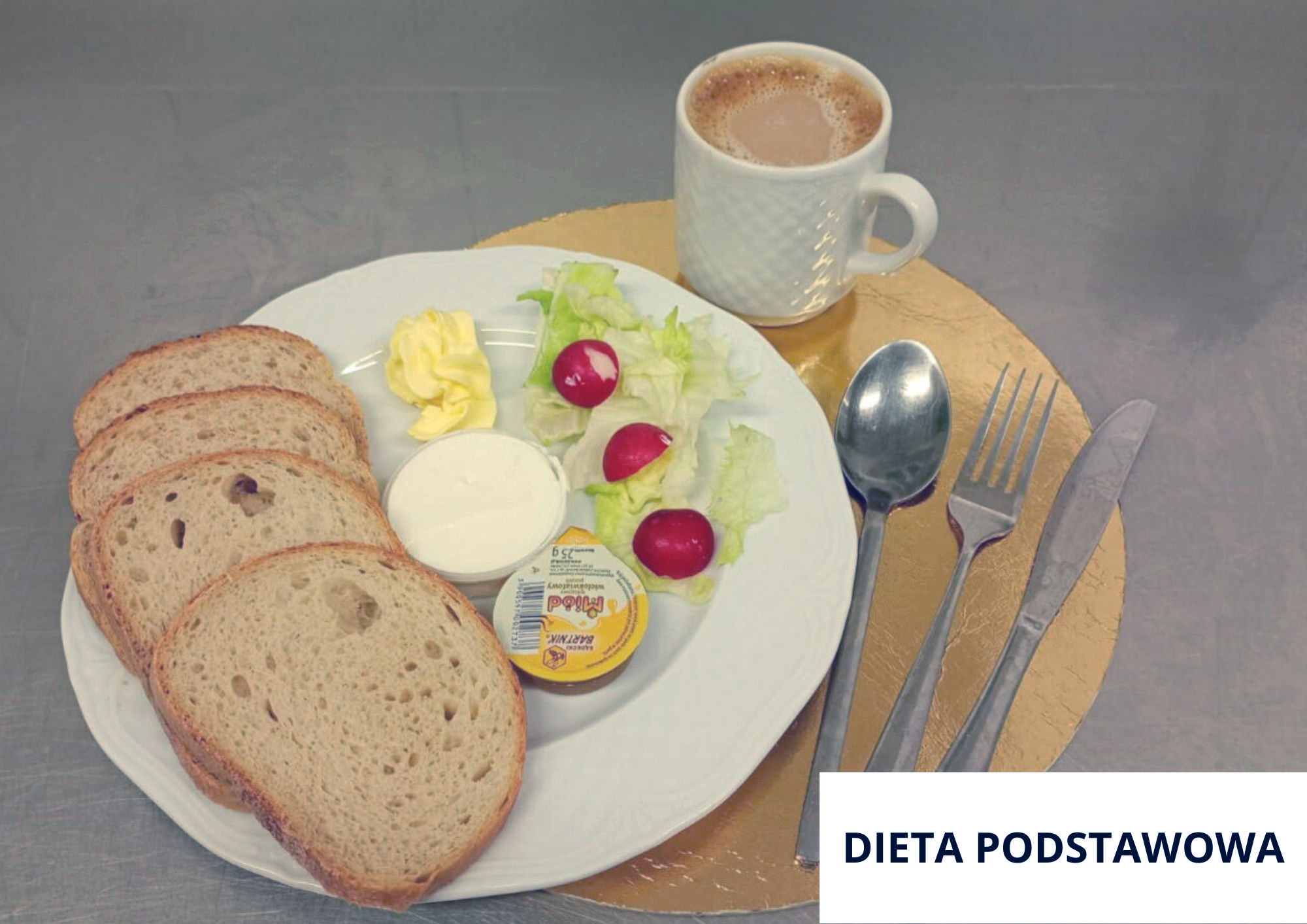 Na stalowym blacie, na okrągłej złotej podkładce ustawiono biały talerz z czterema kromkami pieczywa. Na talerzu znajduje się także mały kubeczek białego nabiału (jogurt lub serek), porcja masła oraz liście sałaty z trzema małymi, czerwonymi rzodkiewkami. Na talerzu leży również mały, okrągły, zamknięty w opakowaniu produkt porcjowany (np. pasztet/ser topiony) z widoczną etykietą. Z tyłu stoi biały kubek z jasnym napojem (kawa z mlekiem/kakao). Po prawej stronie na podkładce ułożone są sztućce: łyżka, nóż i widelec. W prawym dolnym rogu widoczny napis „DIETA PODSTAWOWA”