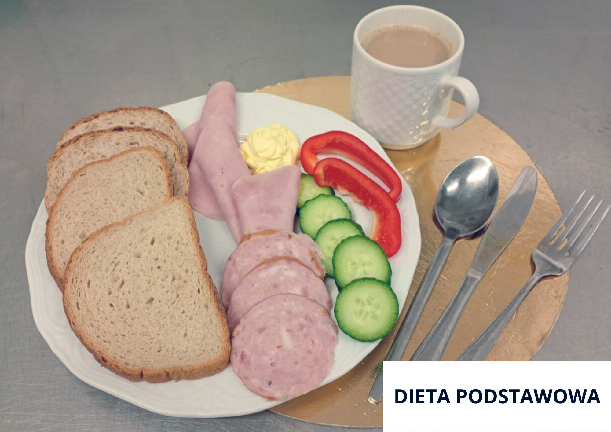 Na stalowym blacie, na złotej okrągłej podkładce znajduje się biały talerz z czterema kromkami pieczywa po lewej stronie. Na środku talerza ułożono kilka plasterków wędliny: część wędliny zwinięta jest w rulon, a część pokrojona w okrągłe plastry. Obok wędliny widać porcję masła, kilka plasterków świeżego ogórka oraz dwa długie paski czerwonej papryki. Z tyłu stoi biały kubek z jasnym napojem (kawa z mlekiem/kakao). Po prawej stronie leżą sztućce: łyżka, nóż i widelec. W prawym dolnym rogu umieszczono napis „DIETA PODSTAWOWA”
