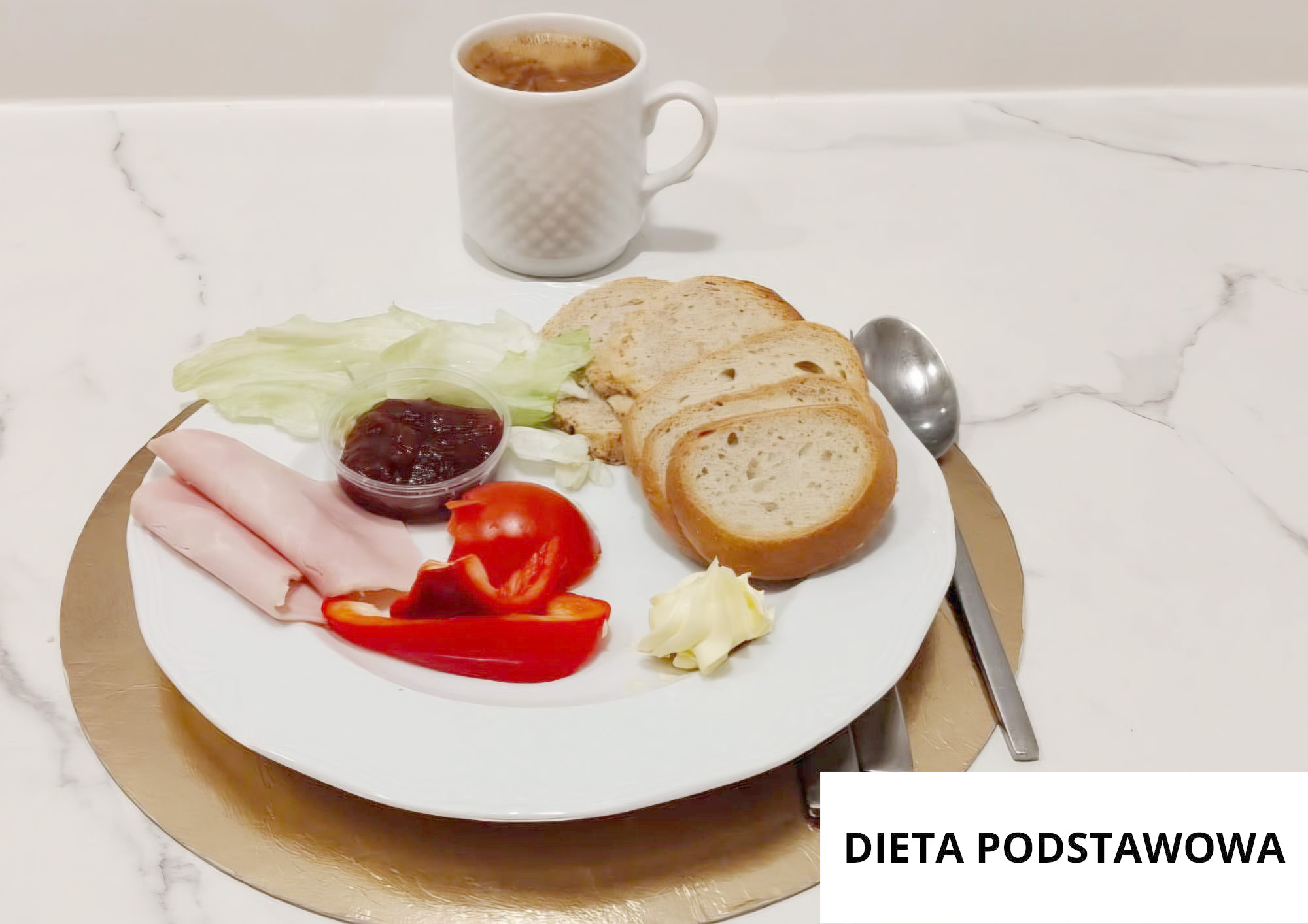 Na jasnym, marmurkowym blacie stoi okrągła złota podkładka z białym talerzem. Na talerzu ułożono trzy kromki pieczywa, kilka cienkich plasterków jasnej wędliny, liście sałaty oraz kawałki czerwonej papryki (połówka i kilka pasków). Obok warzyw stoi mały, przezroczysty pojemniczek z ciemnym dżemem, a z przodu talerza widoczna jest porcja masła w kształcie rozetki. Z tyłu, nad talerzem, stoi biały kubek z ciemnym napojem (herbata lub kawa). Po prawej stronie, częściowo pod talerzem, leżą sztućce: łyżka i nóż. W prawym dolnym rogu widoczna jest biała plansza z napisem „DIETA PODSTAWOWA”