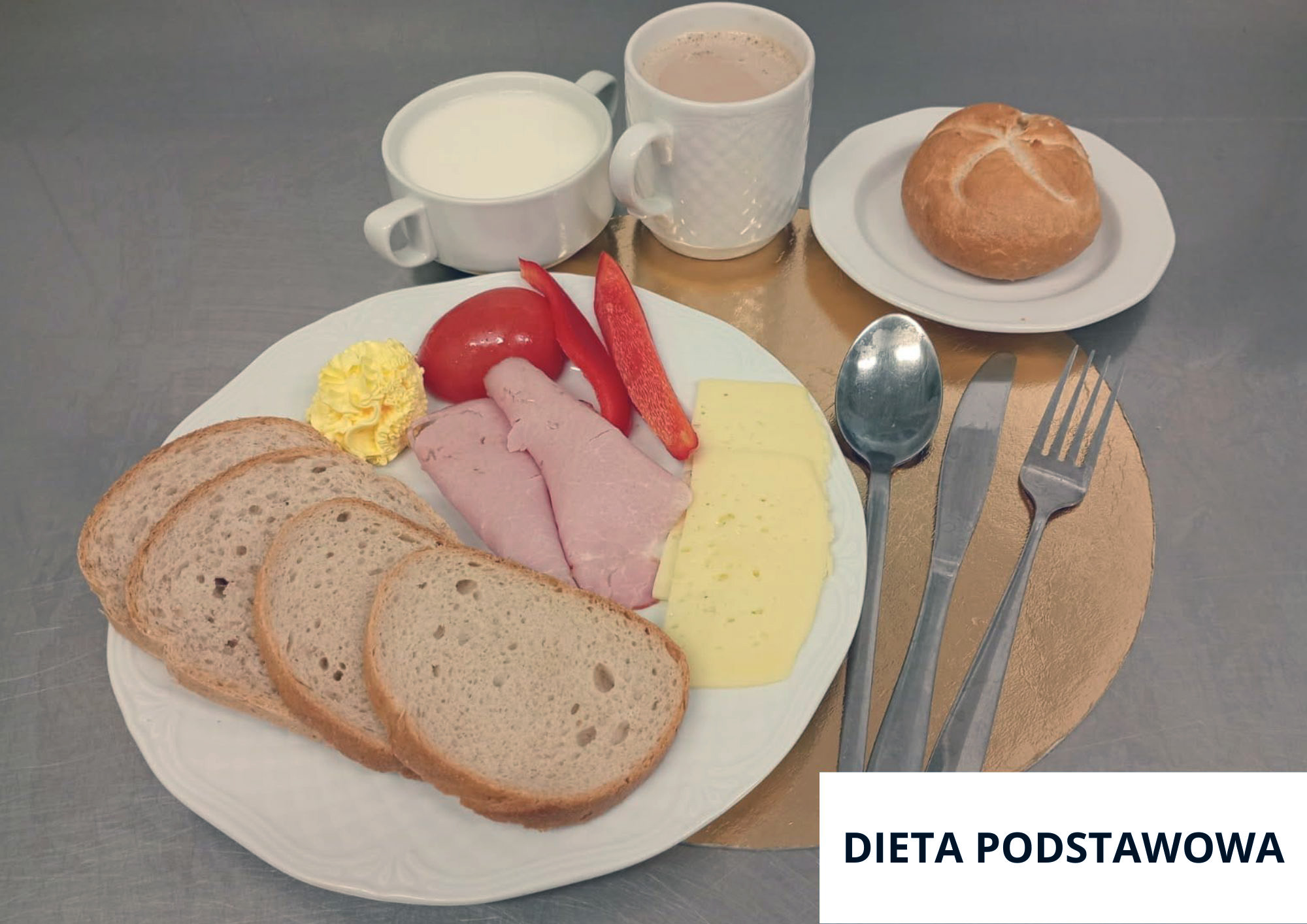 Na talerzu trzy kromki chleba, dwa plasterki wędliny drobiowej, dwa plasterki sera żółtego, porcja masła oraz świeże warzywa: pomidor i czerwona papryka. W tle filiżanka mleka i kubek kawy z mlekiem, obok bułka kajzerka na małym talerzyku. Sztućce ułożone po prawej stronie