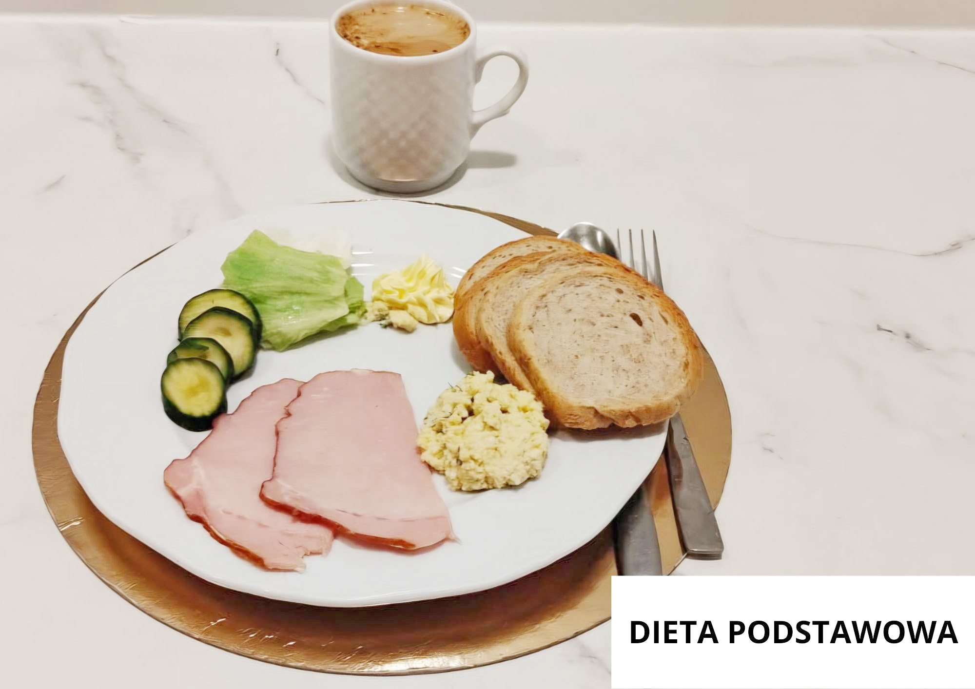 Na białym talerzu trzy plasterki szynki, pasta jajeczna, dwie kromki chleba pszenno-żytniego, liść sałaty oraz plasterki świeżego ogórka. Obok porcja masła. W tle kubek kawy z mlekiem. Talerz ustawiony na złotej podkładce, sztućce po prawej stronie