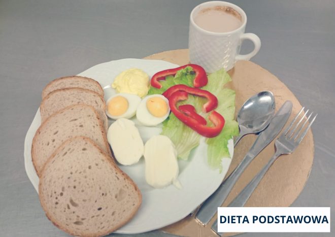 Śniadanie w diecie podstawowej podane na białym talerzu ustawionym na złotej podkładce. Na talerzu znajdują się cztery kromki pieczywa, przekrojone jajko na twardo, porcja masła oraz plasterki czerwonej papryki ułożone na liściach sałaty. Obok widoczne są kawałki białego sera. W tle stoi biały kubek z jasnym napojem. Po prawej stronie talerza ułożone są sztućce: łyżka, nóż i widelec. W prawym dolnym rogu widoczny napis ‘DIETA PODSTAWOWA’