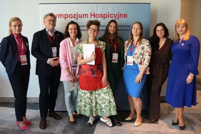 Grupowe zdjęcie organizatorów i uczestników sympozjum hospicyjnego stojących przed banerem wydarzenia. Wśród nich znajduje się lekarz Jacek Graliński oraz przedstawicielki zespołu organizacyjnego