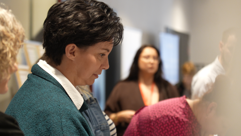 Uczestniczka sympozjum hospicyjnego przegląda materiały konferencyjne podczas części warsztatowej wydarzenia. W tle widoczne są inne osoby biorące udział w spotkaniu