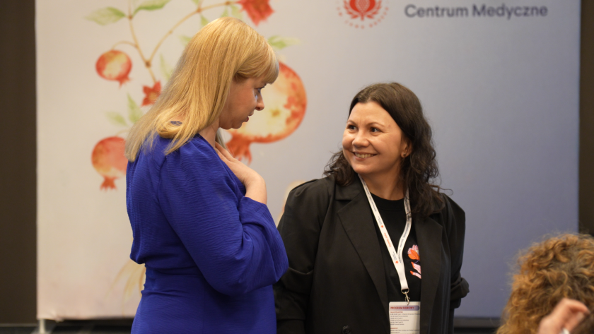 Dwie uczestniczki sympozjum rozmawiają w przerwie między panelami. W tle widoczny jest baner Bonifraterskiego Centrum Medycznego z motywem graficznym granatów