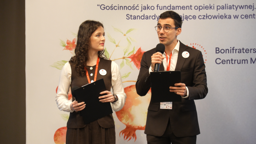 Prowadzący sympozjum – kobieta i mężczyzna – stoją na scenie i zapowiadają kolejną część programu wydarzenia, trzymając mikrofony oraz notatniki