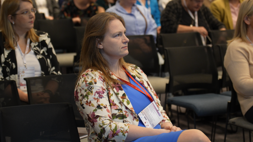 Uczestniczka sympozjum siedzi na sali konferencyjnej i uważnie słucha prelekcji. Na szyi ma identyfikator konferencyjny