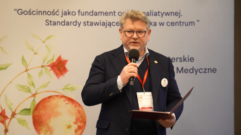 Lekarz Jacek Graliński przemawia do uczestników sympozjum hospicyjnego, trzymając mikrofon i dokument w ręku. W tle widoczny jest baner z hasłem wydarzenia: „Gościnność jako fundament opieki paliatywnej. Standardy stawiające człowieka w centrum”