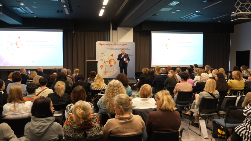 Szerokie ujęcie sali konferencyjnej podczas sympozjum hospicyjnego. Na scenie przemawia prelegent, a uczestnicy wydarzenia siedzą na widowni i słuchają wystąpienia