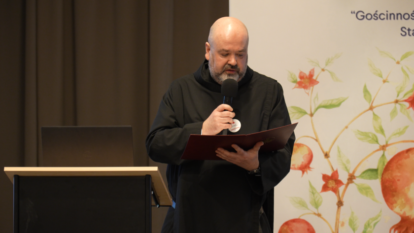 Brat bonifrater w habicie odczytuje tekst z przygotowanych materiałów podczas sympozjum hospicyjnego, stojąc przy mównicy z mikrofonem