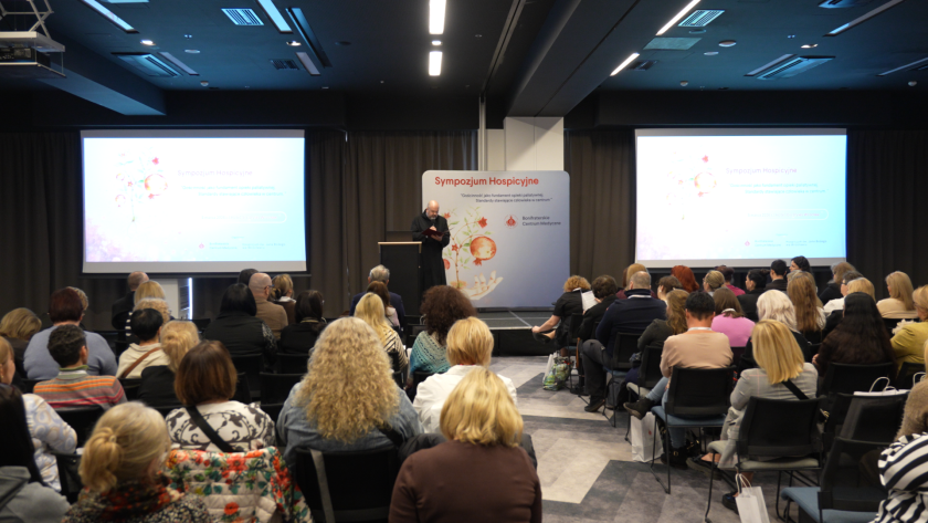 Widok sali konferencyjnej podczas sympozjum hospicyjnego. Na scenie brat bonifrater czyta przygotowane wystąpienie, a uczestnicy siedzą na widowni i słuchają prelekcji