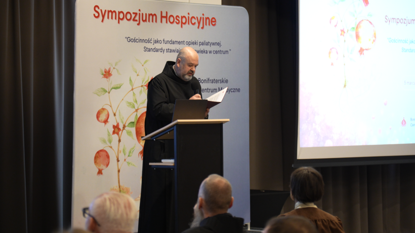 Brat bonifrater stoi przy mównicy i odczytuje tekst z dokumentu podczas sympozjum hospicyjnego. W tle widoczny jest baner wydarzenia z logo Bonifraterskiego Centrum Medycznego