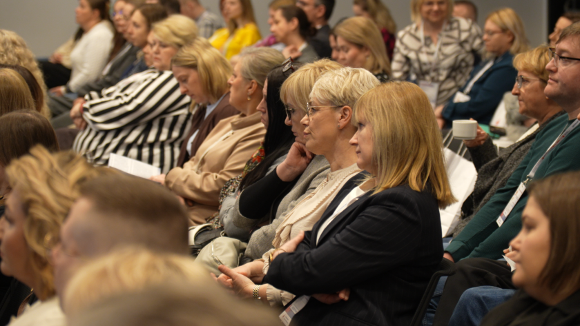 Uczestnicy sympozjum siedzą na sali konferencyjnej i uważnie słuchają wystąpienia. W pierwszych rzędach widoczne są osoby z materiałami konferencyjnymi i identyfikatorami