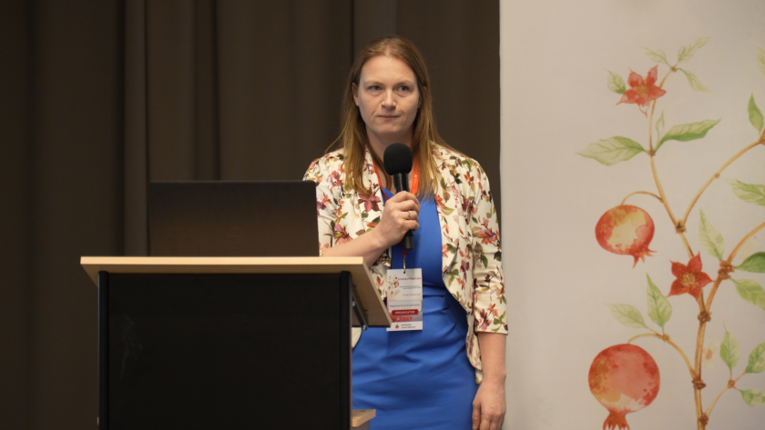 Prelegentka stoi przy mównicy z mikrofonem i prowadzi wystąpienie podczas sympozjum hospicyjnego. Obok znajduje się laptop wykorzystywany do prezentacji