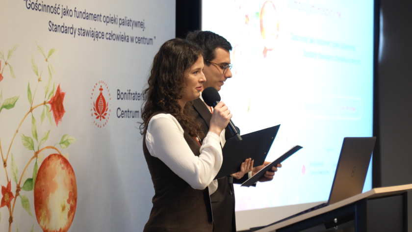 Prowadzący sympozjum – kobieta i mężczyzna – stoją na scenie przy mównicy i zapowiadają kolejną część programu wydarzenia, trzymając mikrofony oraz notatniki. W tle widoczny jest baner Bonifraterskiego Centrum Medycznego