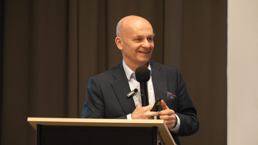 Prelegent stoi przy mównicy i przemawia do uczestników sympozjum hospicyjnego, trzymając mikrofon i pilot do prezentacji