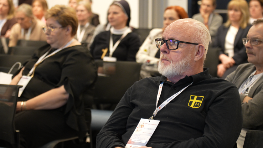 Uczestnik sympozjum siedzi na sali konferencyjnej i uważnie słucha wykładu. Na szyi ma identyfikator konferencyjny