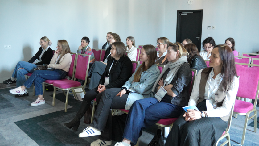 Grupa uczestników bierze udział w warsztatach odbywających się w mniejszej sali. Siedzą na krzesłach ustawionych w rzędach i słuchają prowadzącej