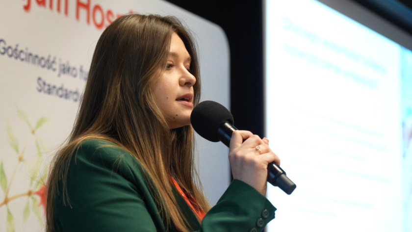 Prelegentka przemawia do uczestników sympozjum, trzymając mikrofon. W tle widoczny jest baner wydarzenia dotyczącego opieki hospicyjnej