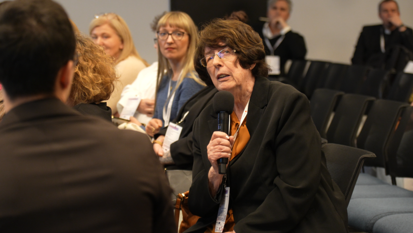 Uczestniczka sympozjum siedzi na sali konferencyjnej i zadaje pytanie, trzymając mikrofon w ręku. W tle widoczni są inni uczestnicy wydarzenia