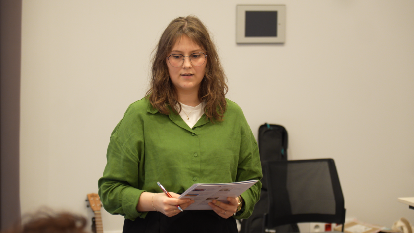 Prelegentka stoi na scenie i odczytuje tekst z materiałów konferencyjnych, trzymając kartkę oraz długopis w dłoni