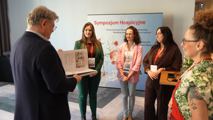 Mężczyzna wręcza podziękowania organizatorkom sympozjum hospicyjnego. Kobiety stoją przed banerem wydarzenia „Sympozjum Hospicyjne – Gościnność jako fundament opieki paliatywnej. Standardy stawiające człowieka w centrum”