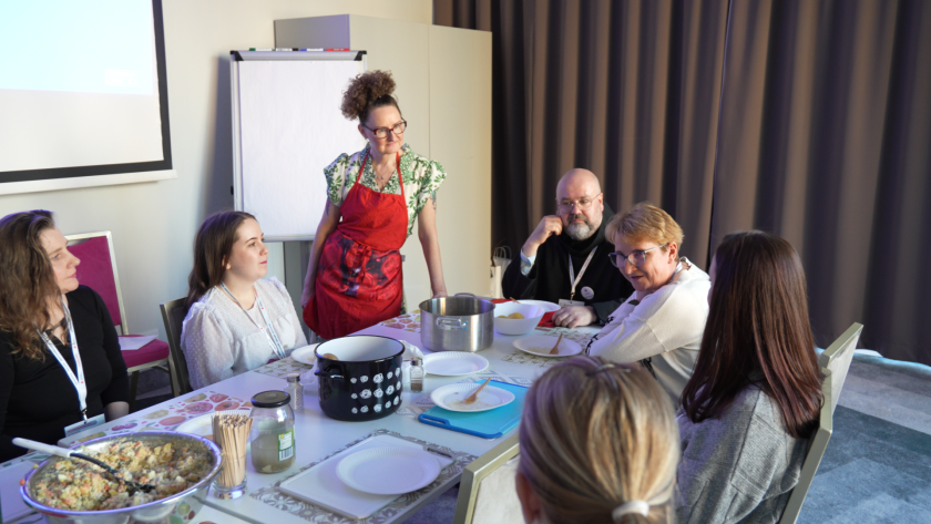 Uczestnicy warsztatów siedzą przy wspólnym stole podczas zajęć towarzyszących sympozjum hospicyjnemu. Prowadząca w czerwonym fartuchu omawia kolejne etapy przygotowania potrawy, a na stole znajdują się garnki, talerze i składniki