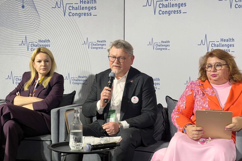 Panel dyskusyjny podczas XI Kongresu Wyzwań Zdrowotnych; jeden z uczestników mówi do mikrofonu, obok siedzą pozostali paneliści, a w tle widoczna jest ściana z logo kongresu