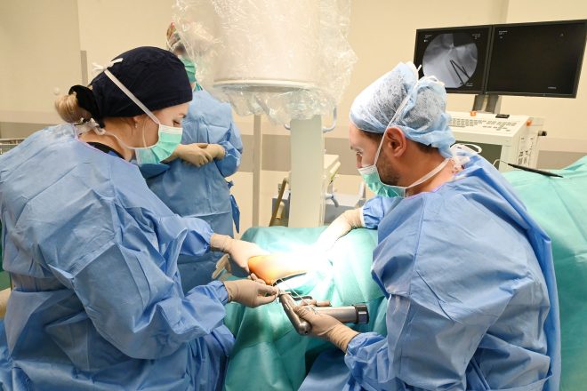 Zespół chirurgiczny w trakcie operacji ortopedycznej stopy – lekarze w sterylnych strojach wykonują zabieg z użyciem specjalistycznego sprzętu, w tle widoczne monitory RTG